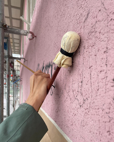 Schriftenmalerin am malen einer Fassadenbeschriftungen von Hand mit Pinsel, Malstock und antrazith Farbe auf rosa Putz.